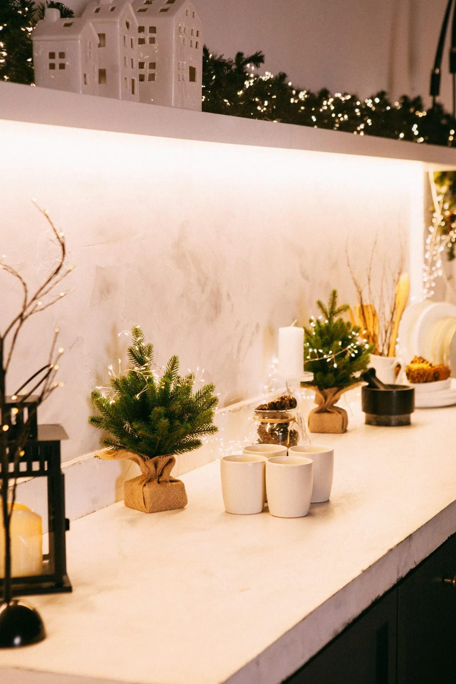 Small Christmas trees in burlap pots lined up on a white counter under warm under-cabinet lights