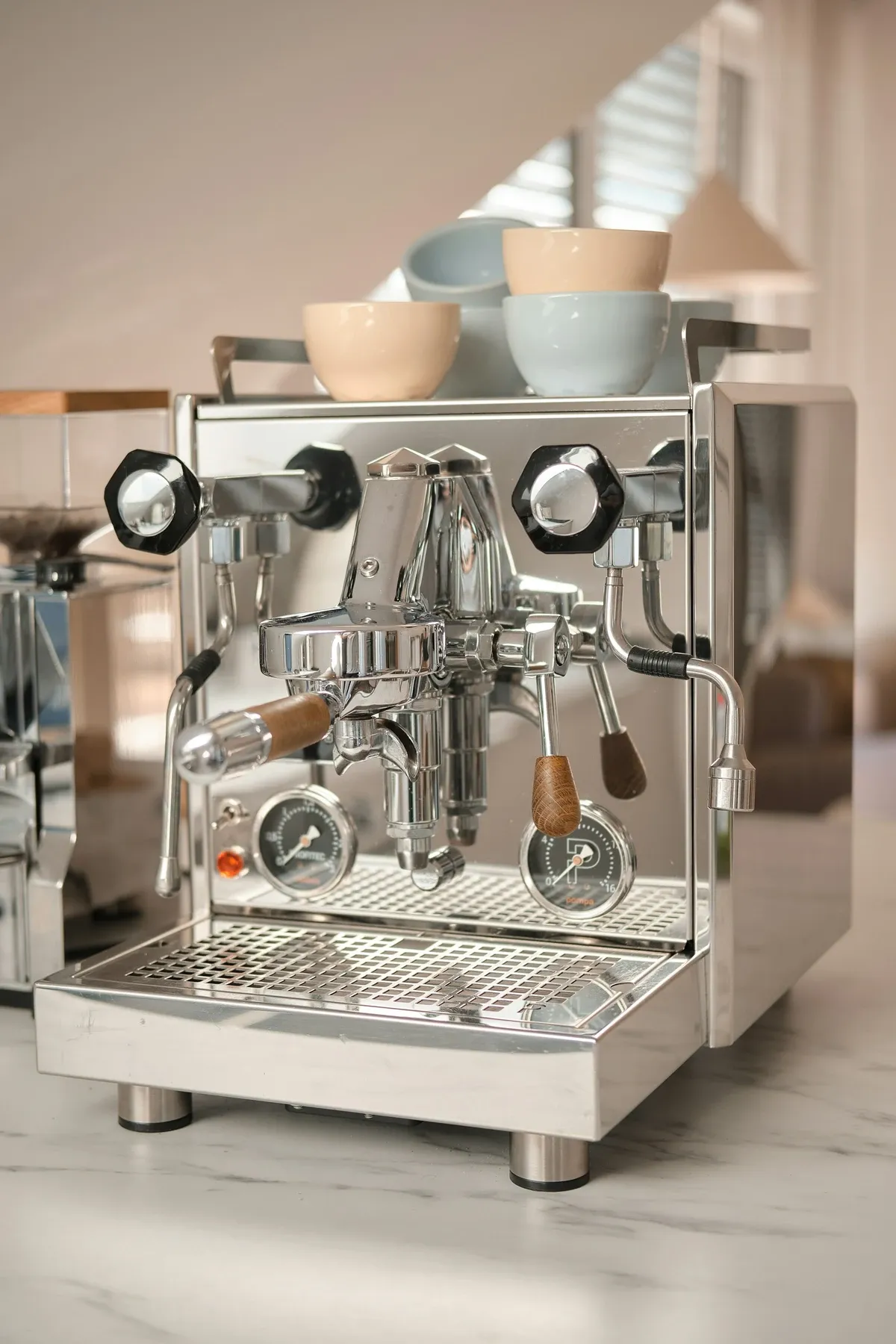 Chrome espresso machine with wood handles and cups warming on top, on a marble counter in soft window light
