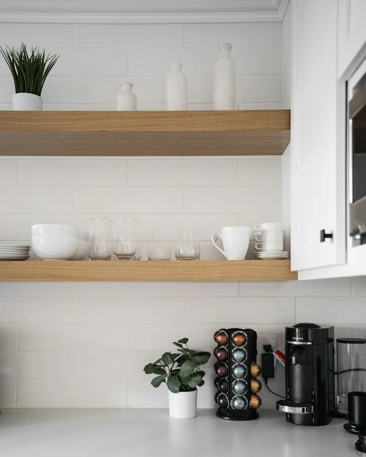 Two floating wood shelves with cups and jars above a coffee station in a white kitchen