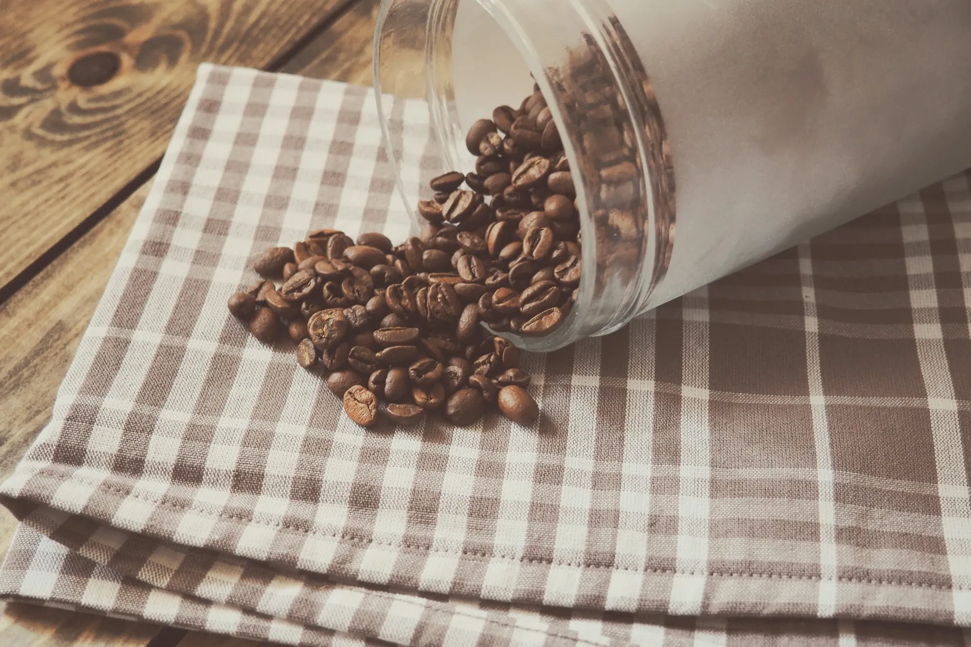 Glass jar tipped on its side spilling roasted coffee beans onto a checked linen napkin on a wood table