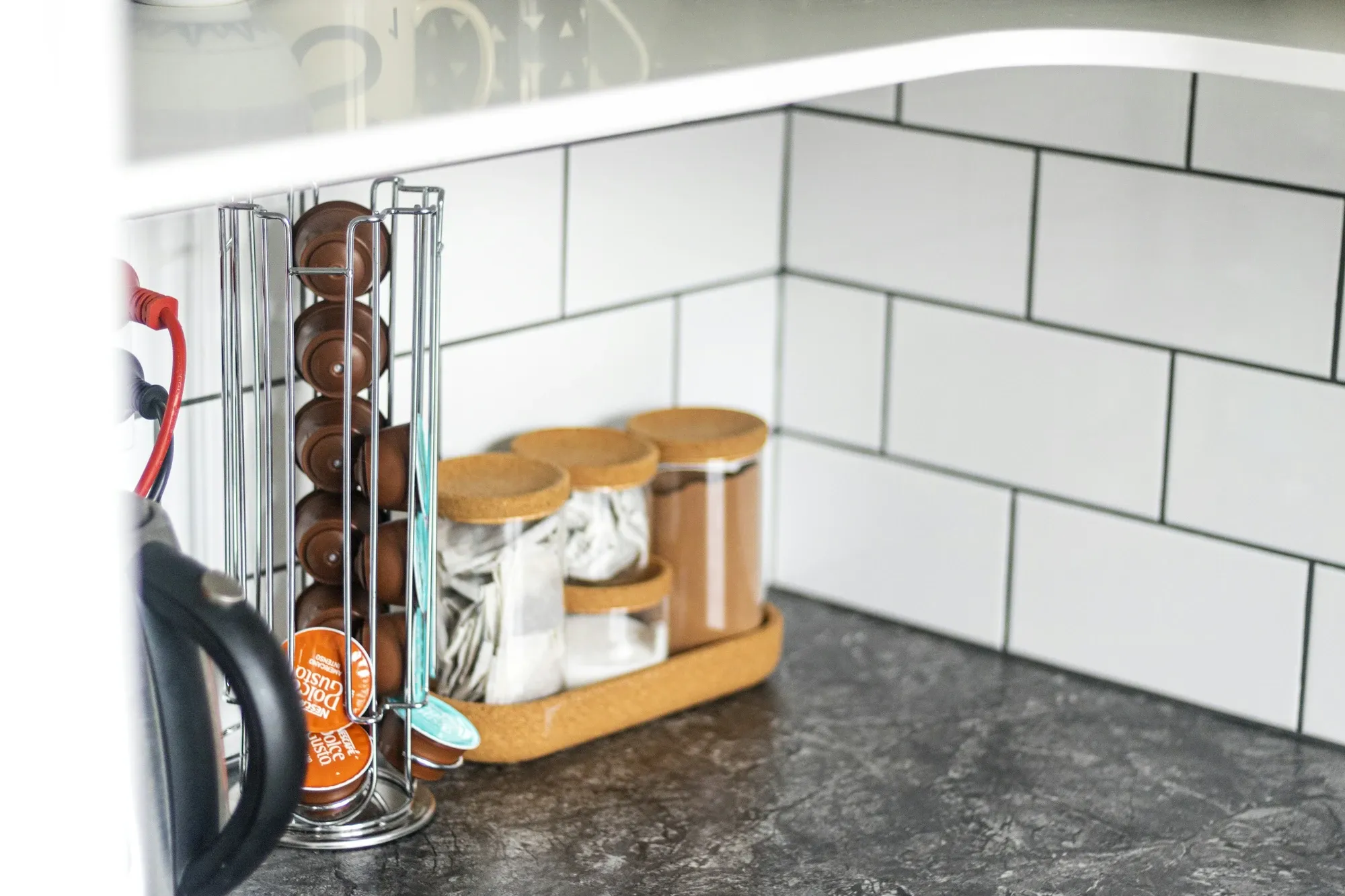 Chrome capsule tower filled with coffee pods beside glass jars with cork lids on a cork tray