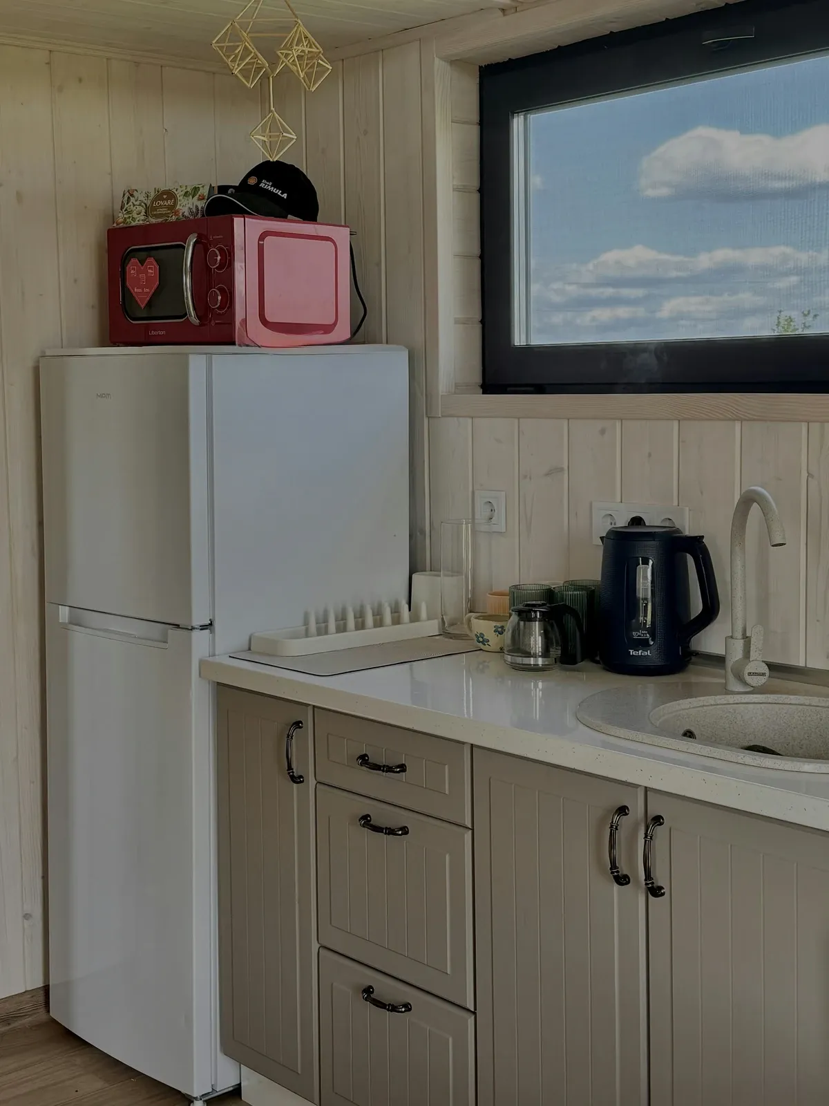 Small kitchen corner with a white fridge, a pink microwave on top and a kettle on the counter beside it