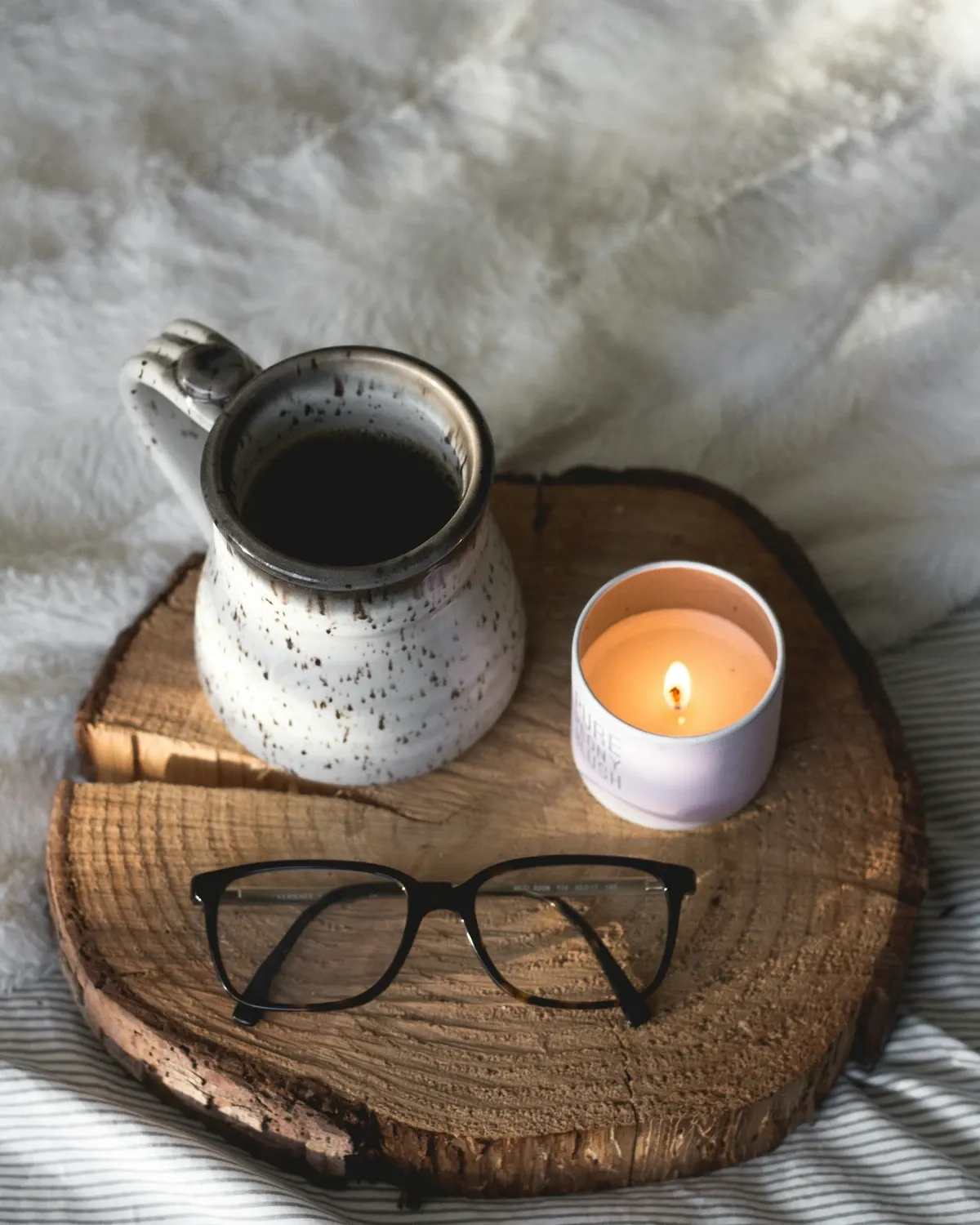 Speckled mug of coffee and a lit candle on a round wood slice tray with eyeglasses on a cream blanket