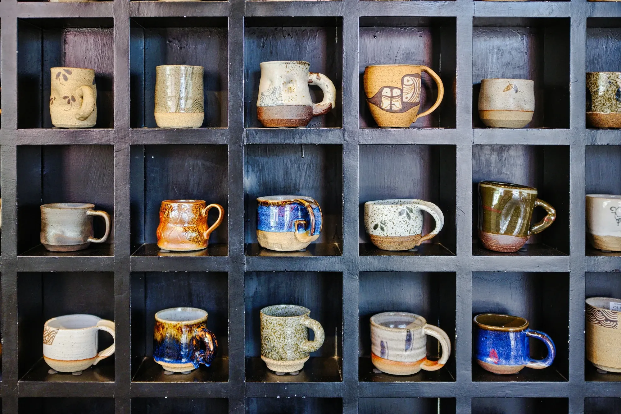 Handmade ceramic mugs in blue, cream and rust glazes, each in its own square cubby of a dark wood wall shelf