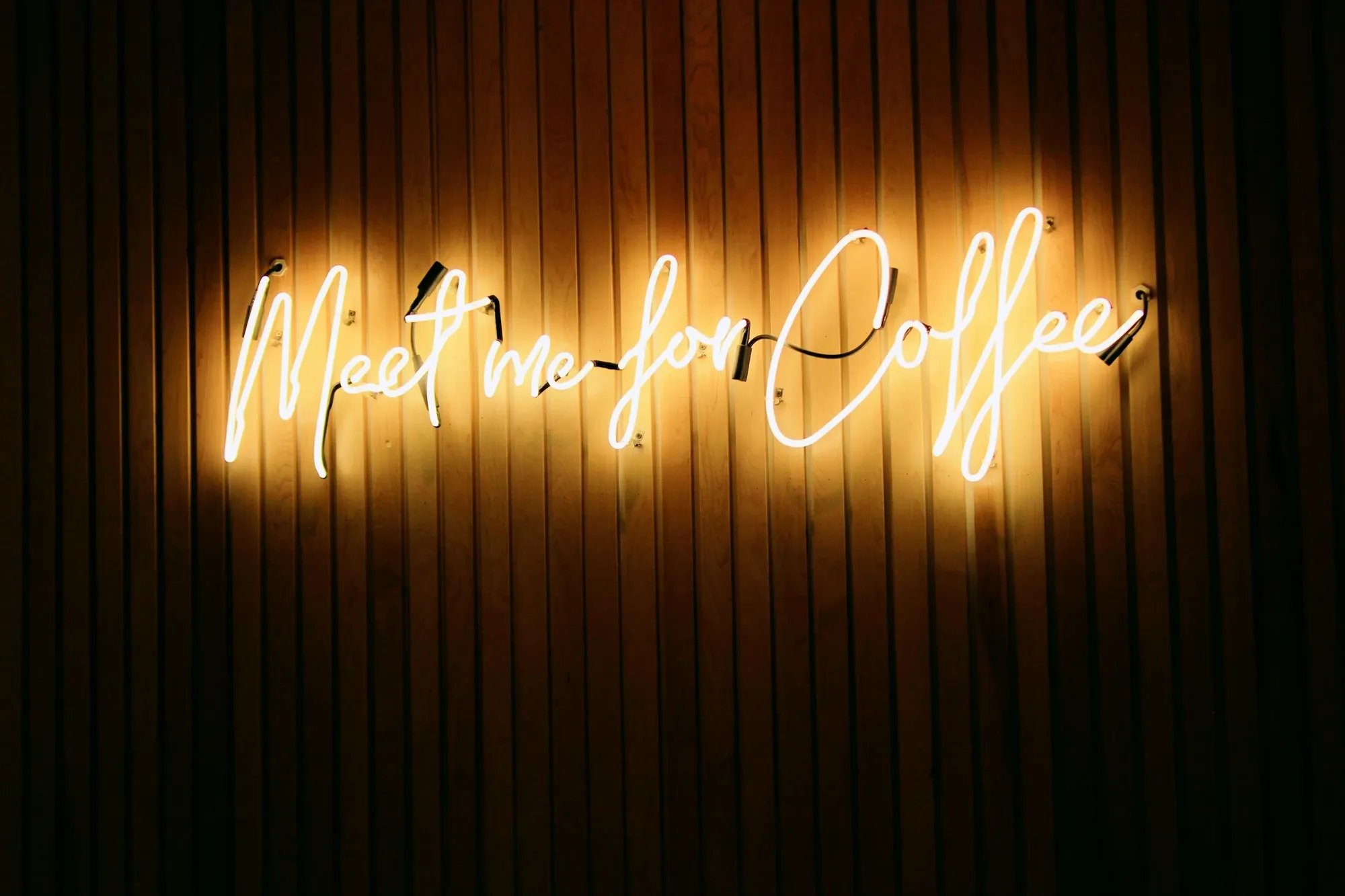 Warm white neon script sign reading Meet me for coffee on a dark vertical wood-slat wall