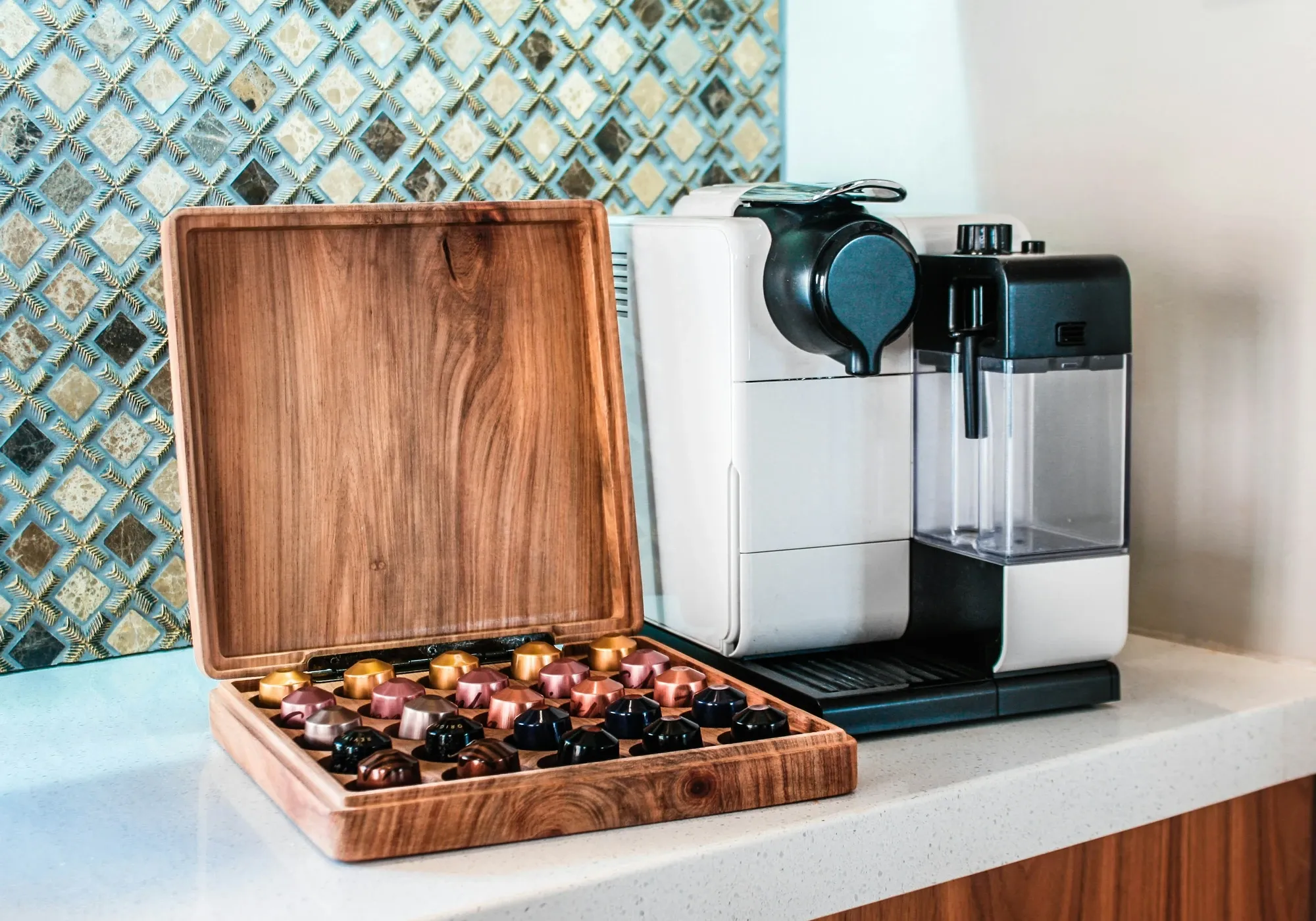 Open wooden box of coffee capsules next to a white capsule espresso machine on a quartz counter with a mosaic backsplash