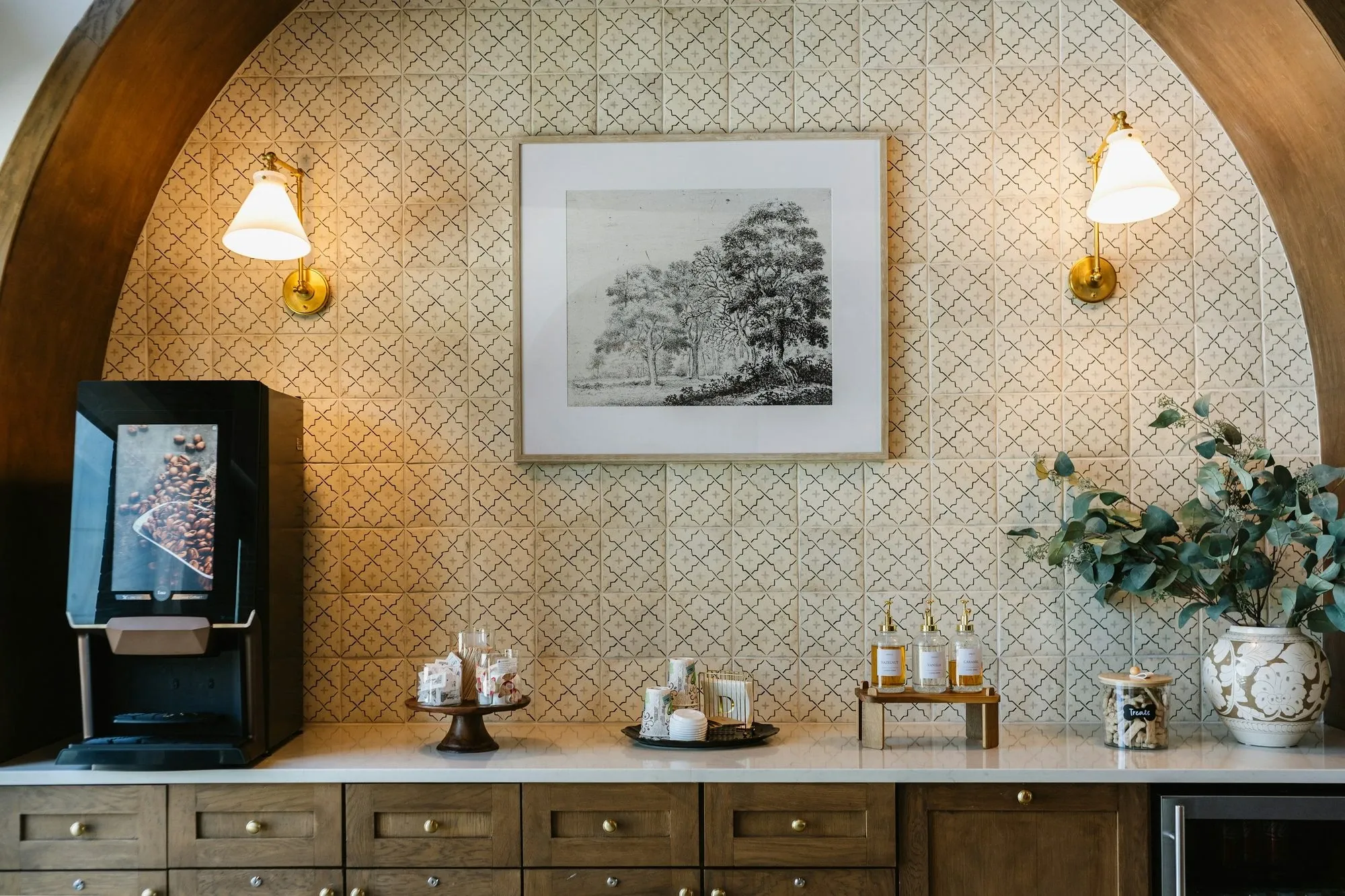 Labeled syrup pump bottles on a small wooden riser at a coffee station under a tiled arch with a framed print