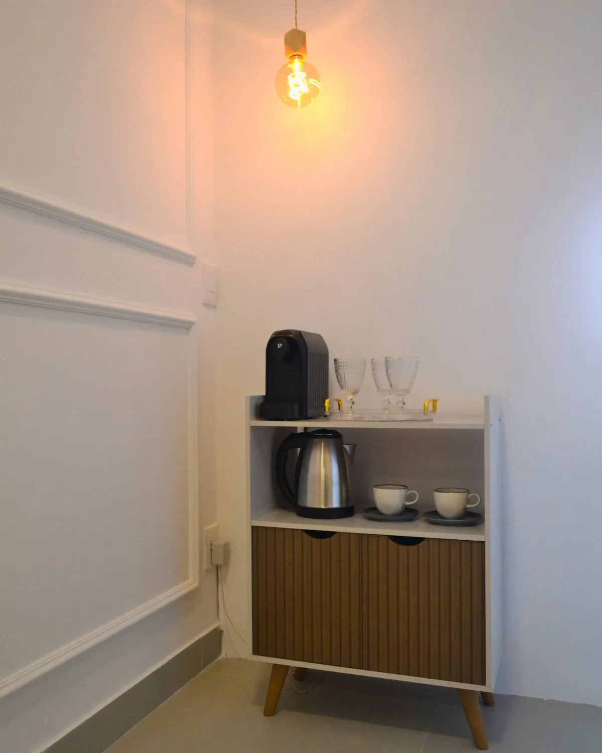 Small white cabinet with a wood front holding a kettle, a pod coffee maker and cups against a white wall