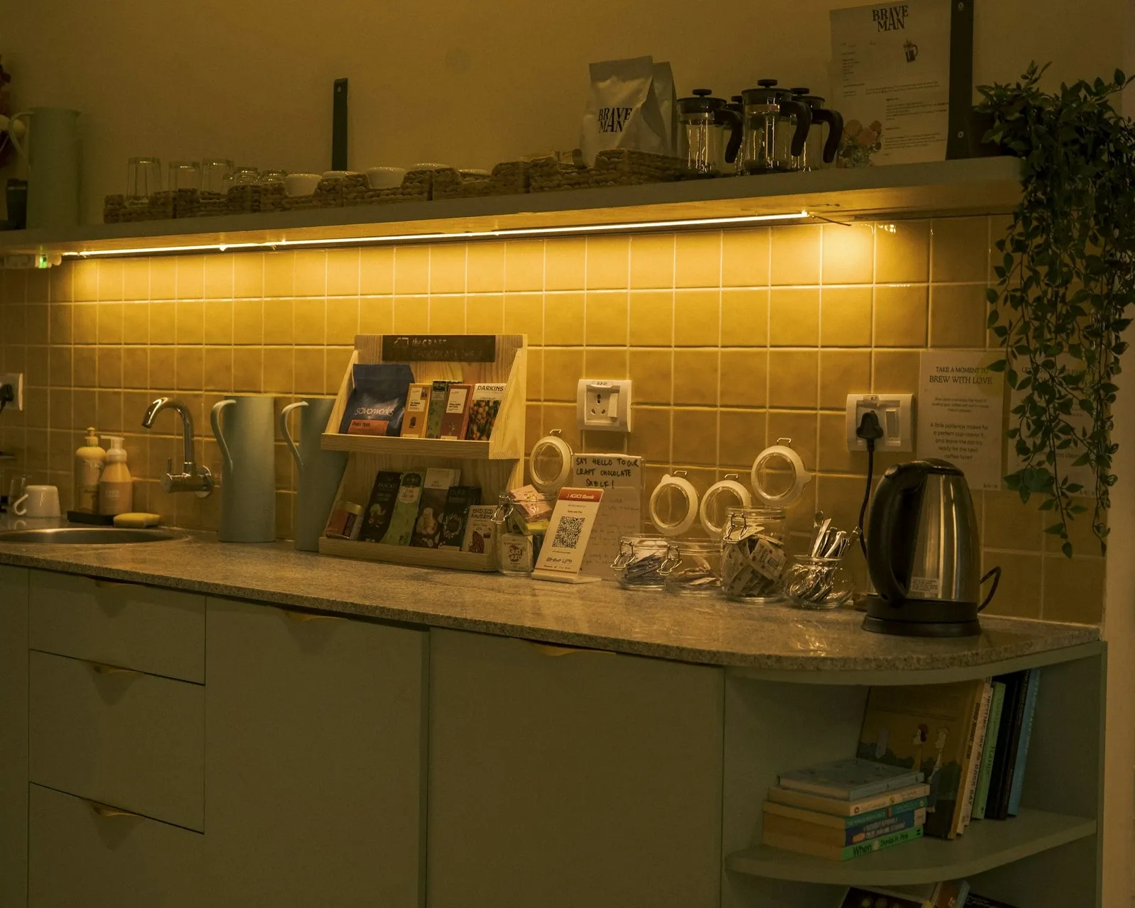 Kitchen counter with tile backsplash lit by warm under-cabinet lights, holding a kettle, mugs and tea supplies