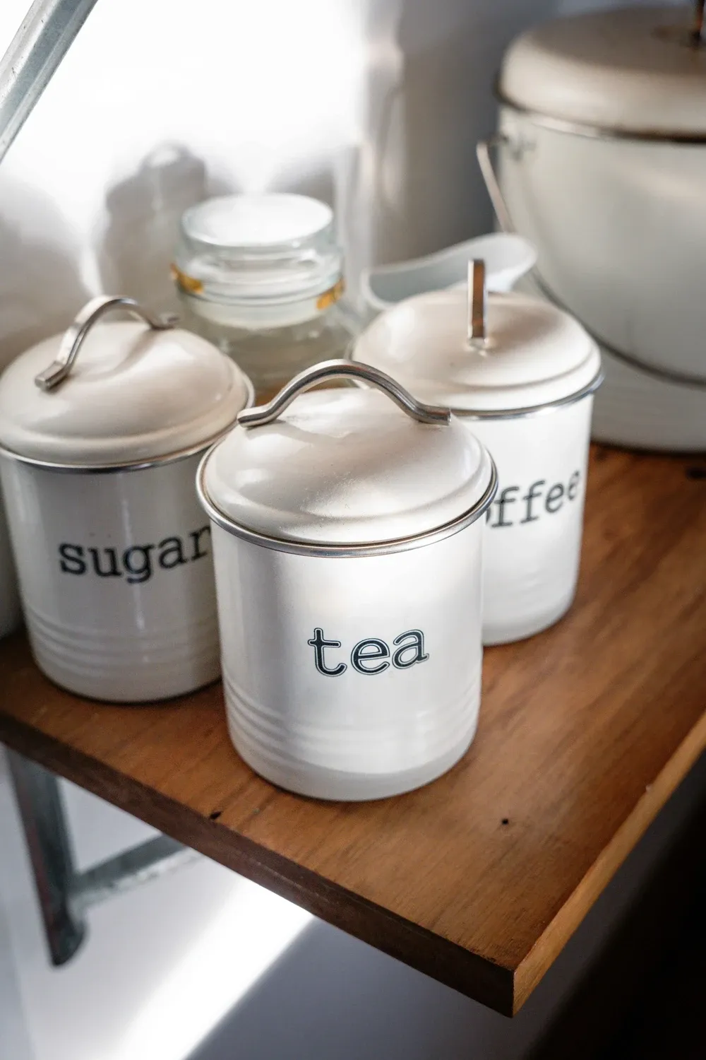 Close view of white enamel canisters labeled sugar, tea and coffee on a wooden shelf