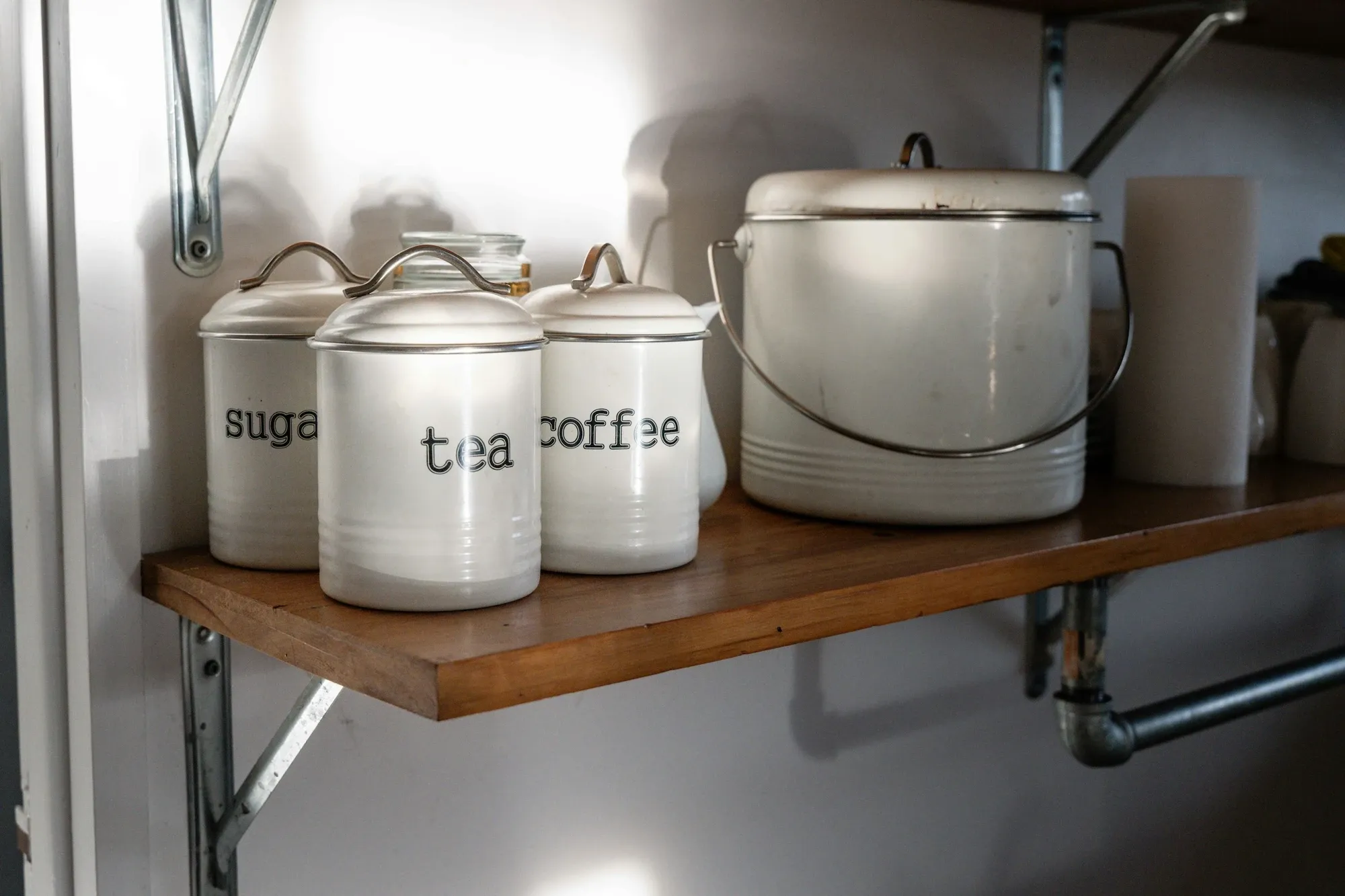 White enamel canisters labeled sugar, tea and coffee in black script on a wooden wall shelf beside a larger enamel pot