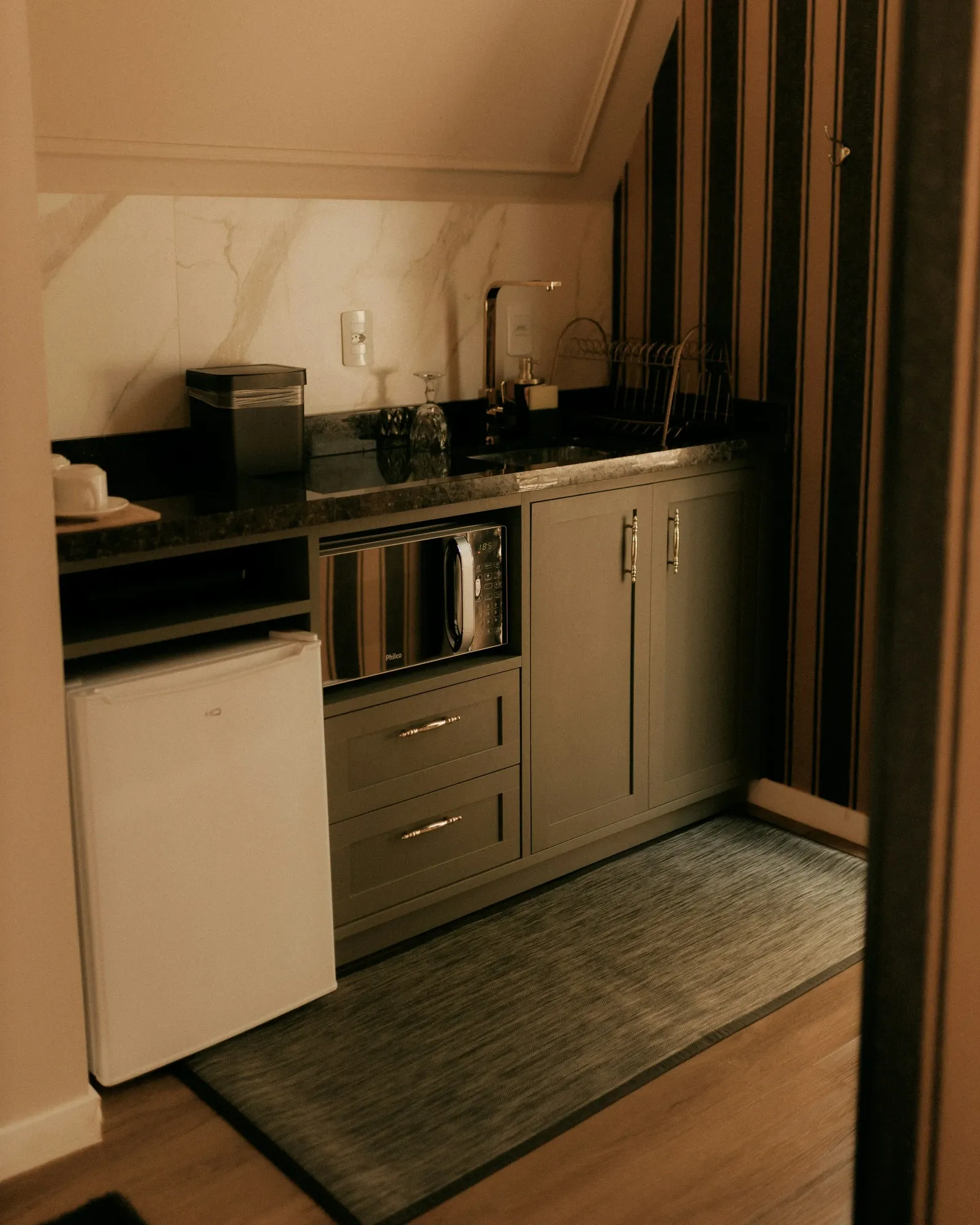 Compact kitchenette under a sloped ceiling with a white mini fridge under the counter beside gray drawers and a dark countertop