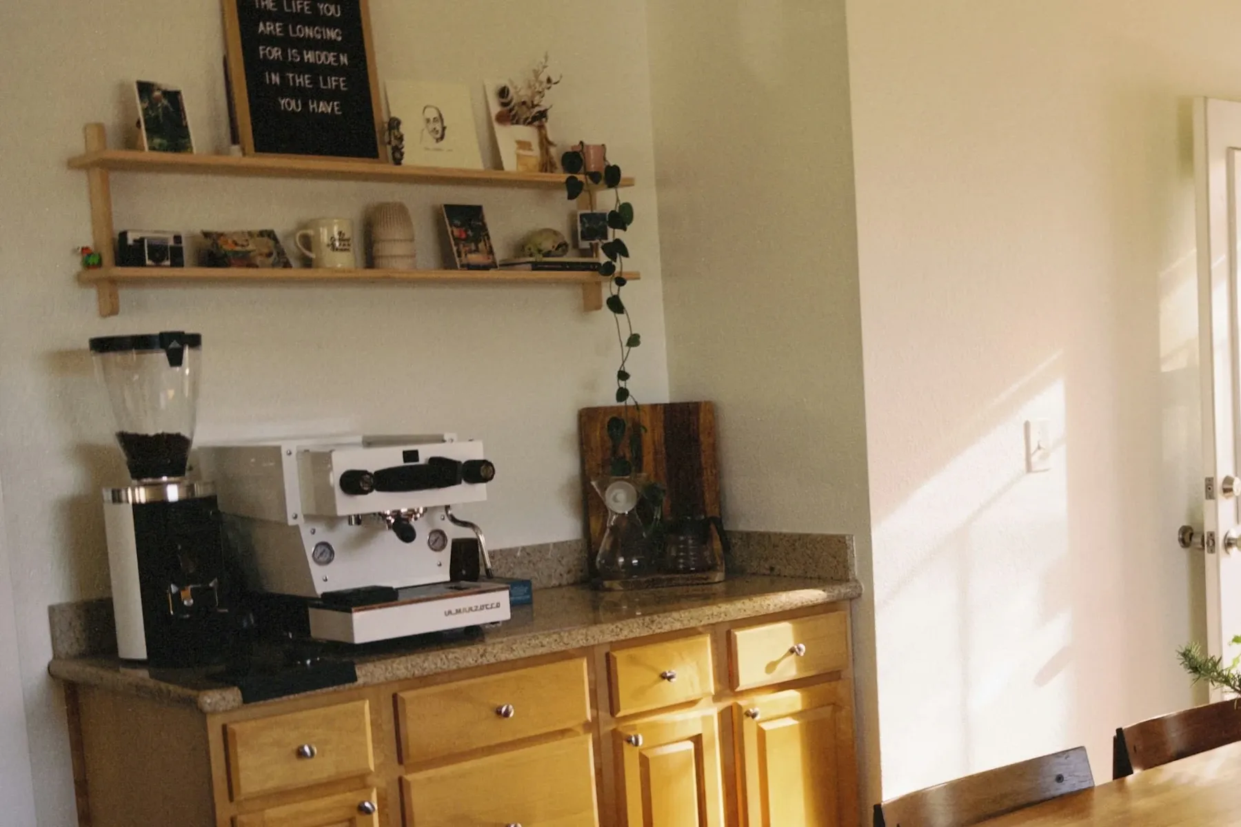 Countertop coffee station with an espresso machine and grinder on a granite counter, two floating shelves of mugs and a letter board above