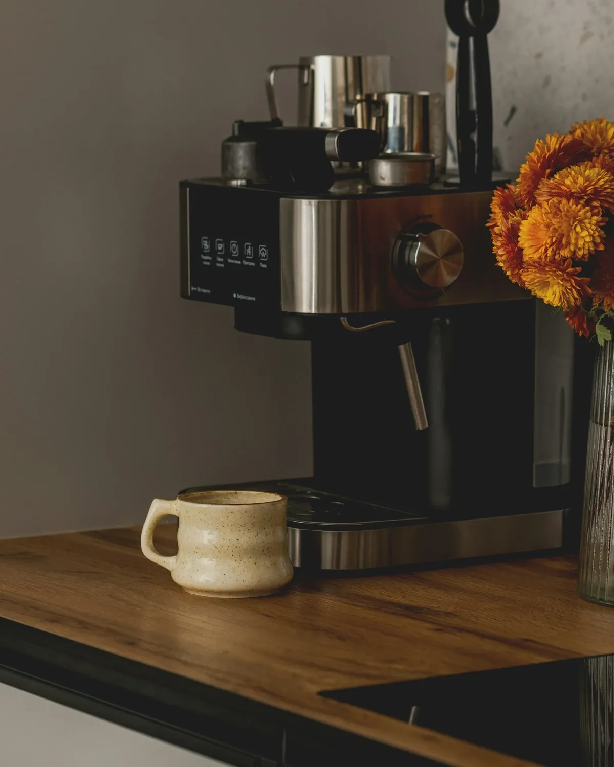 Espresso machine on a wood counter with a cream mug and a vase of orange flowers beside it