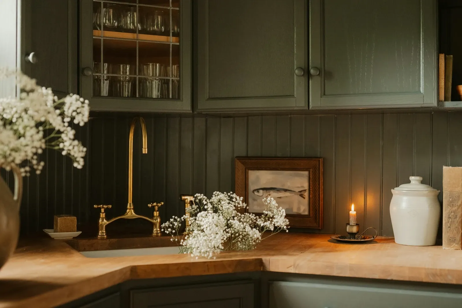 Dark green cabinets and paneled backsplash with a brass faucet, butcher-block counter, white flowers, a framed print and a lit candle