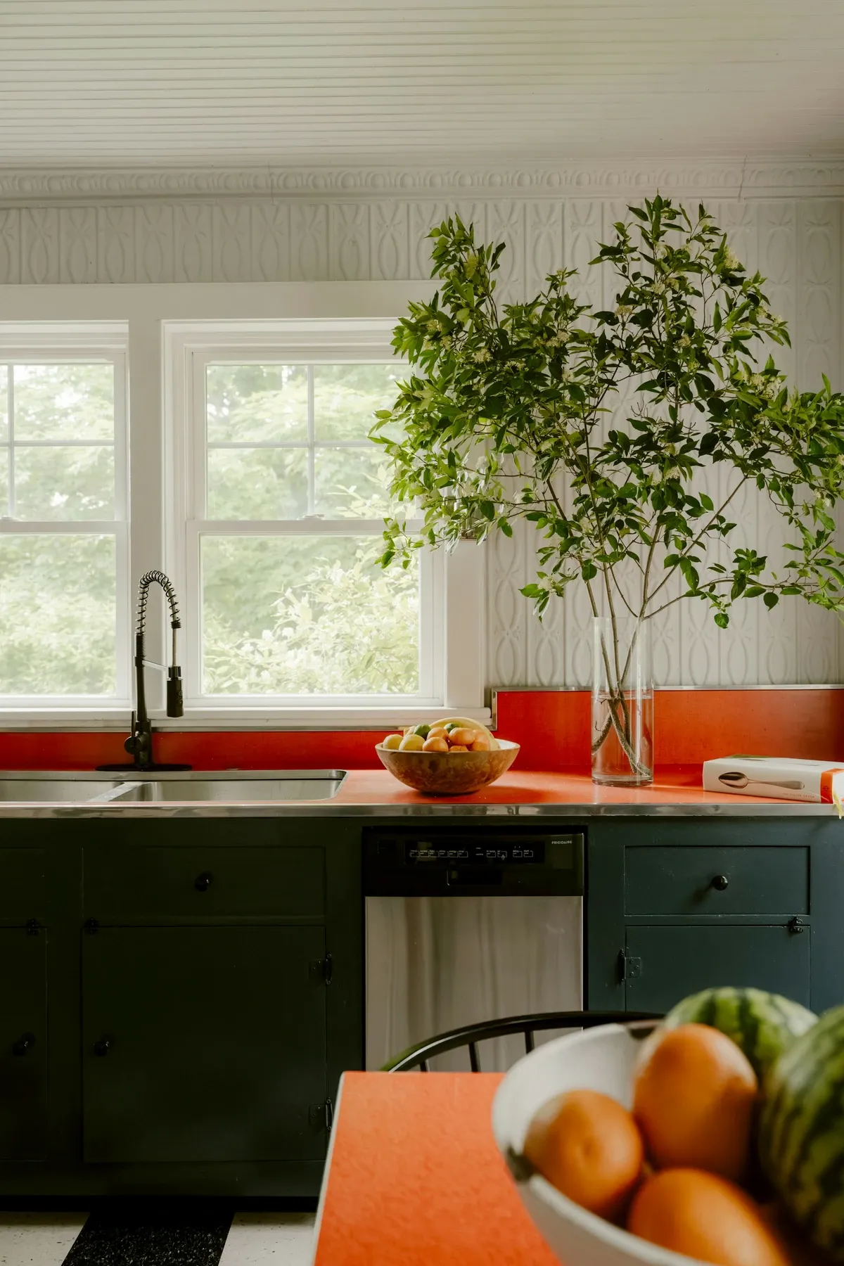 Dark green lower cabinets under a bright orange counter and backsplash by a window, with a potted tree and a bowl of oranges