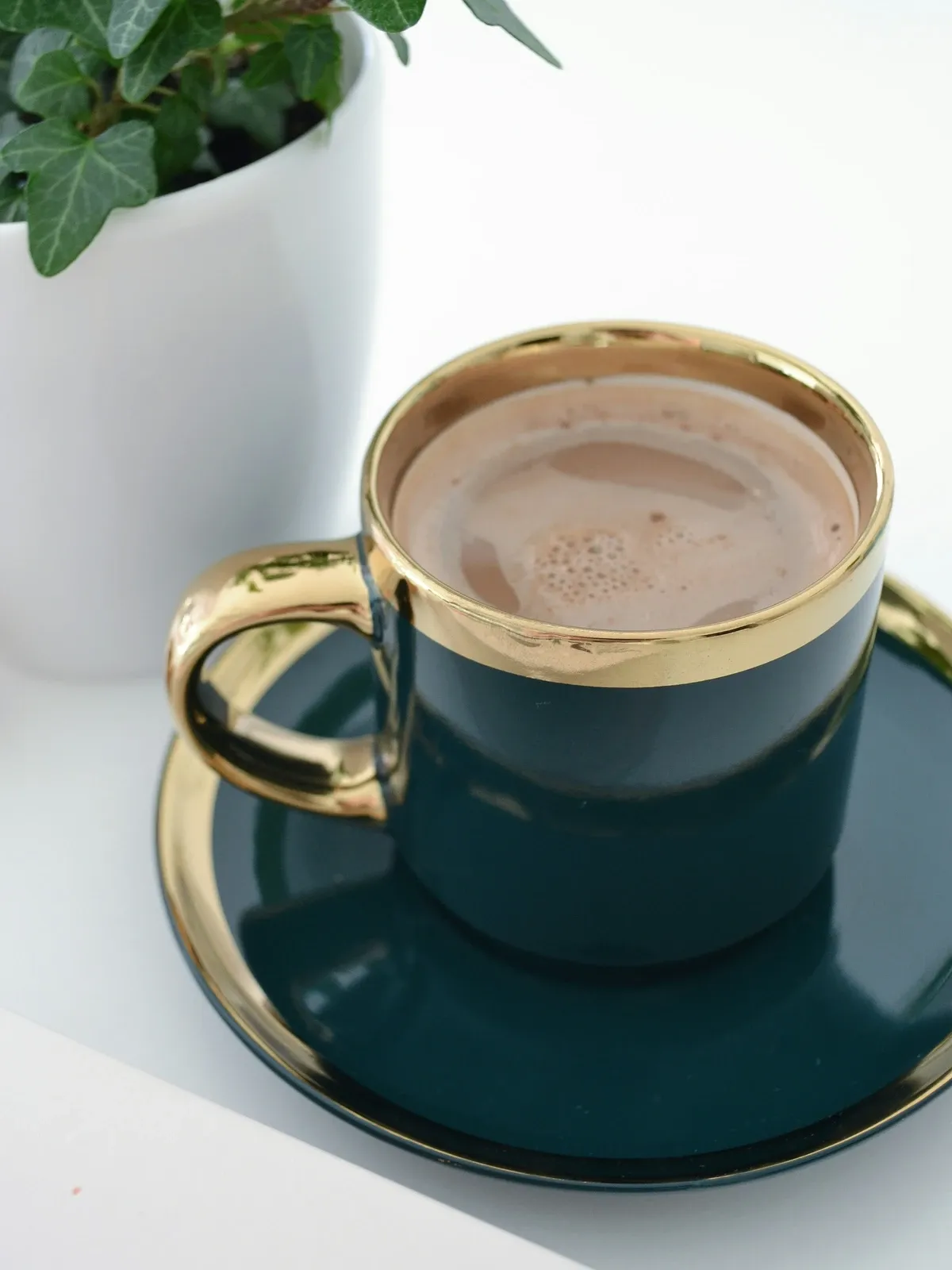 Dark green cup with a wide gold rim on a matching saucer, filled with hot chocolate, beside a white pot with a green plant