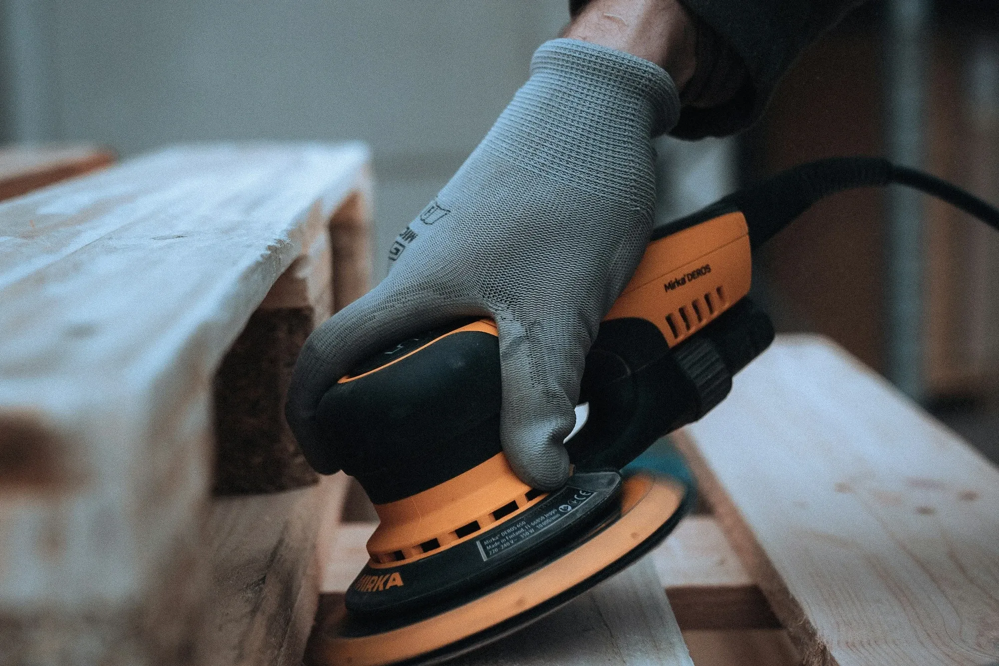 Gloved hand running an orbital sander along a pallet board, the first step of a DIY coffee bar build