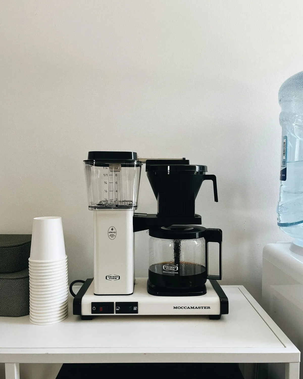 Cream drip coffee maker beside a stack of white paper cups on a small white counter