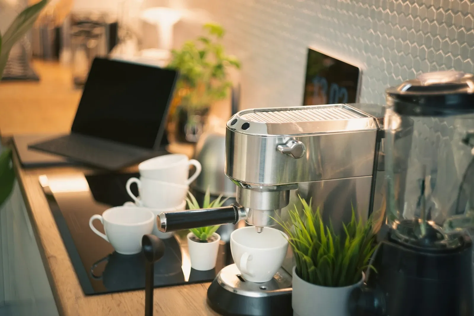 Compact stainless espresso machine with white cups on top, a plant and a laptop on a dark counter