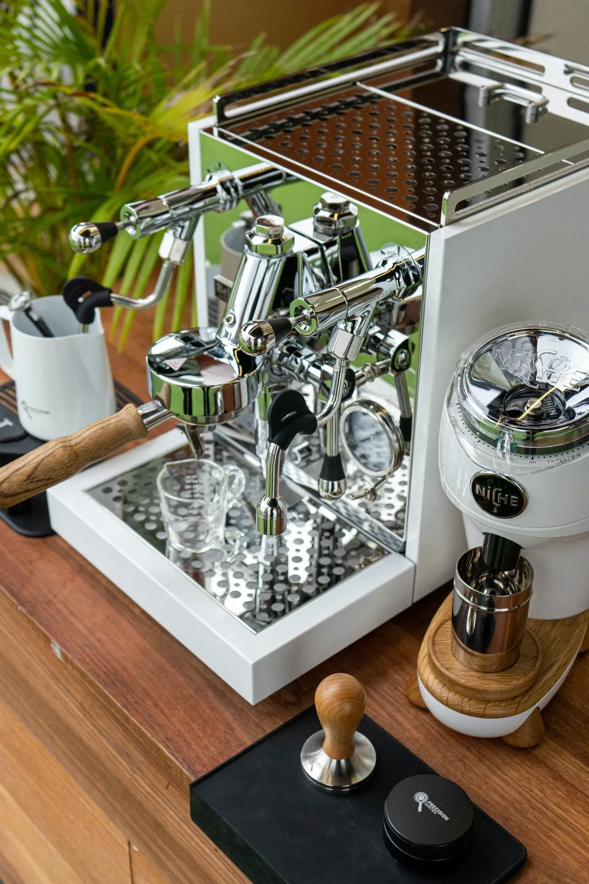Chrome and white espresso machine and grinder on a walnut counter with a wood tamper, a black mat and a green plant behind