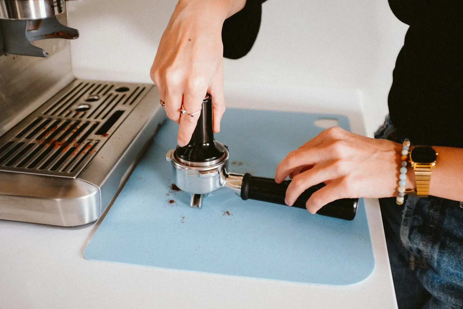 Barista tamping espresso grounds in a portafilter on a blue silicone mat at the counter edge beside an espresso machine