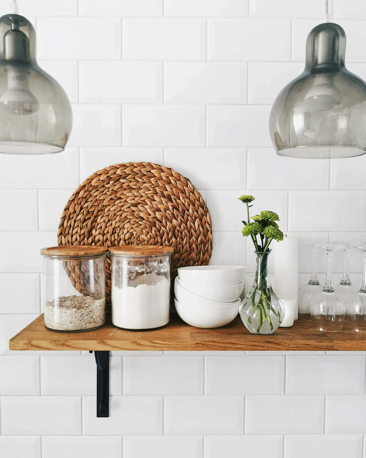 Wood floating shelf with two glass jars, a woven placemat, white bowls and a bud vase against white subway tile