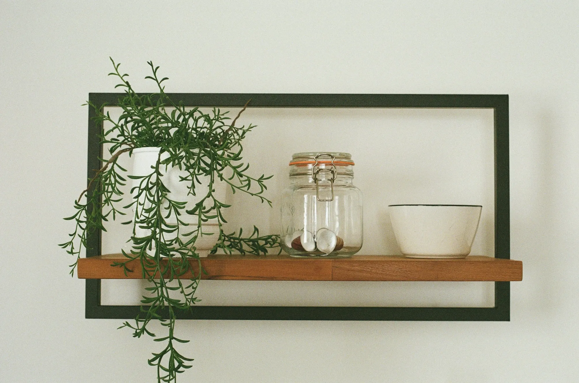 Black-framed wall shelf with a trailing green plant, a glass jar and a white bowl on a white wall