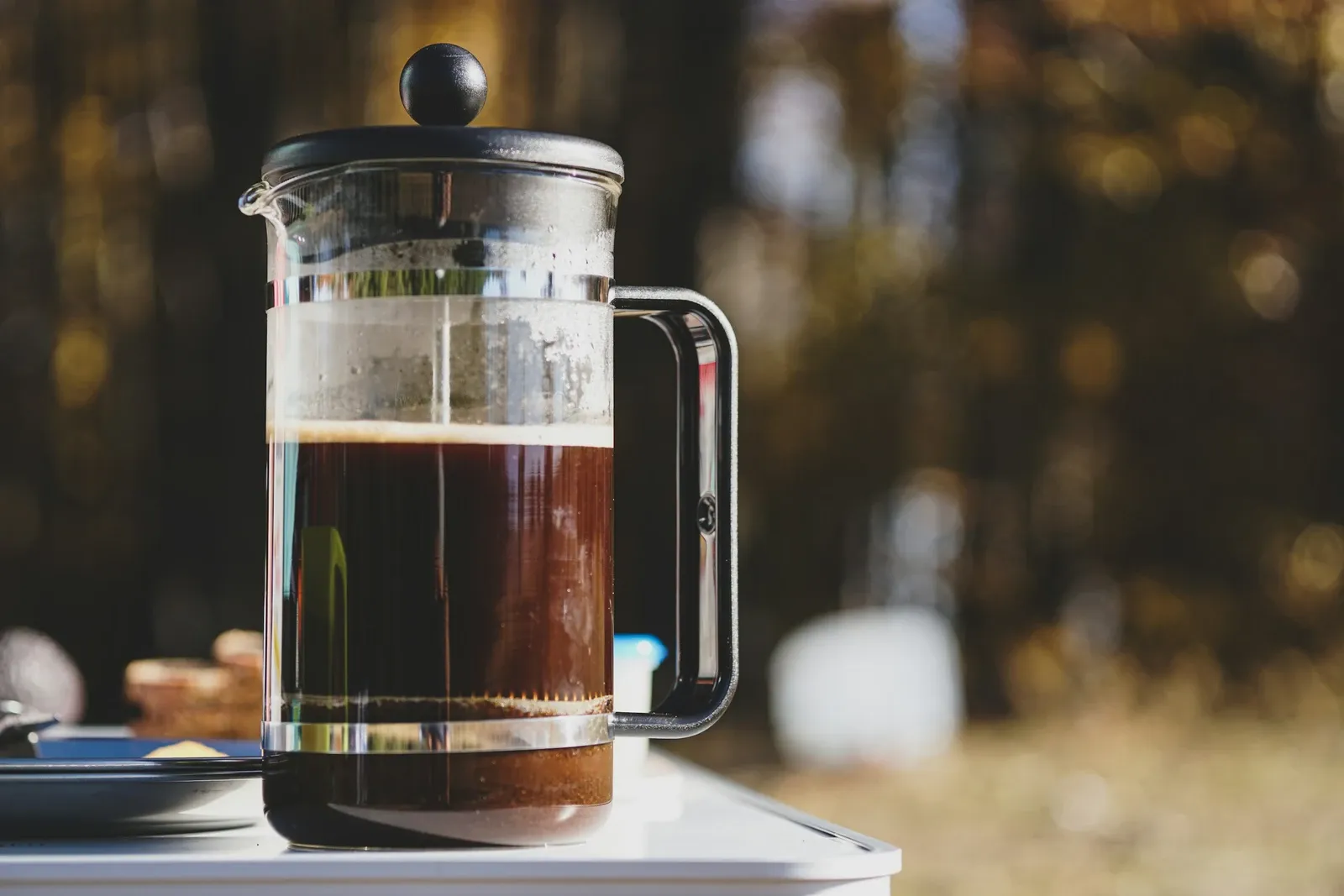 Glass French press full of brewed coffee on a table with a blurred garden behind it