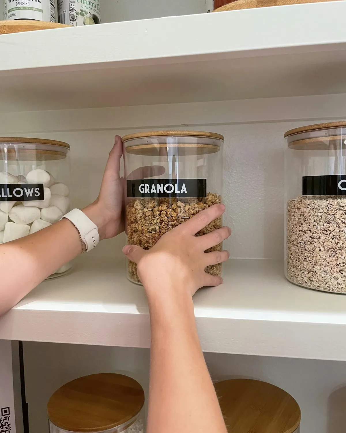 Hands placing a glass jar labeled granola on a white shelf between other labeled glass jars