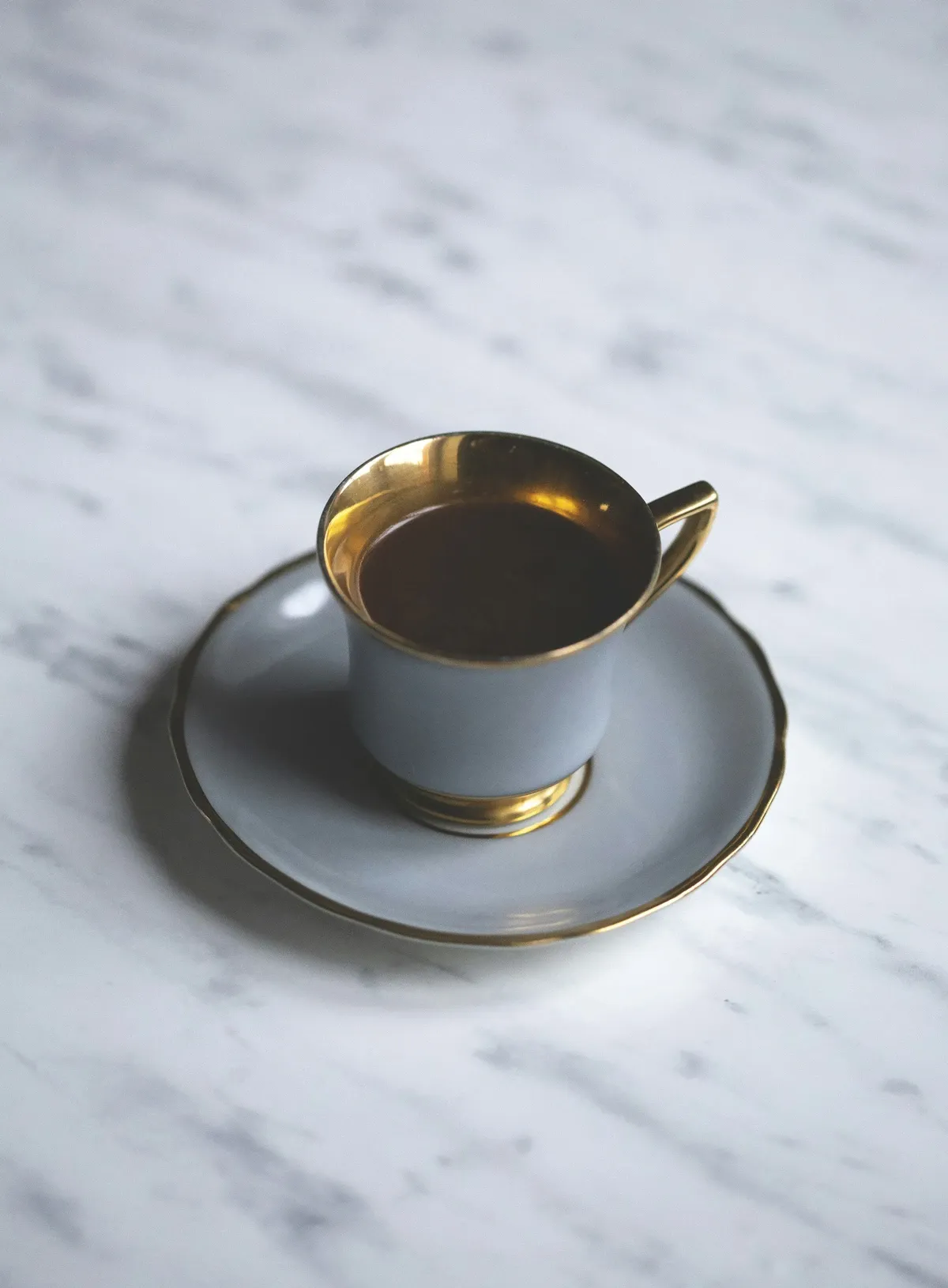 Gray porcelain cup and saucer with a gold rim holding black coffee on a white marble surface