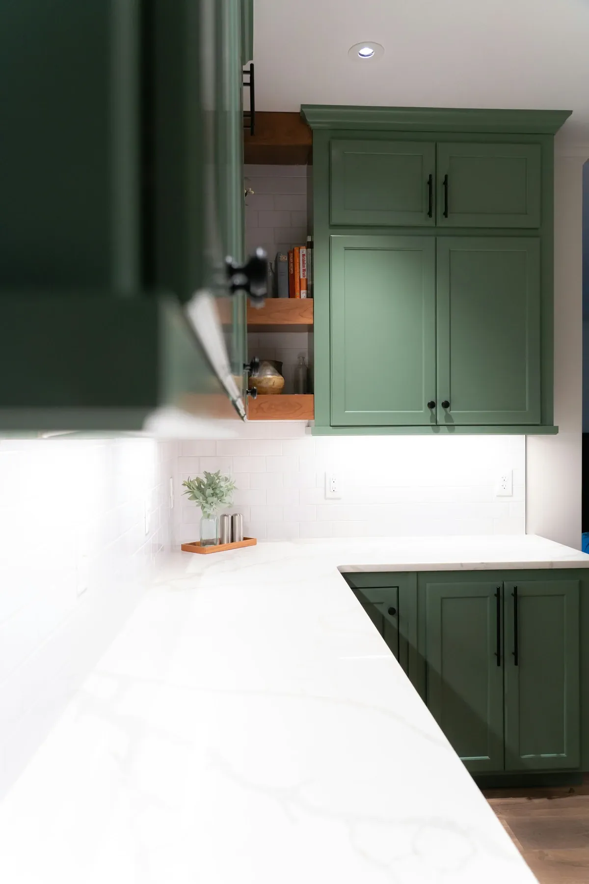 Green shaker cabinets with black pulls and a corner of open wood shelves above a white marble counter, lit subway tile and a small plant tray