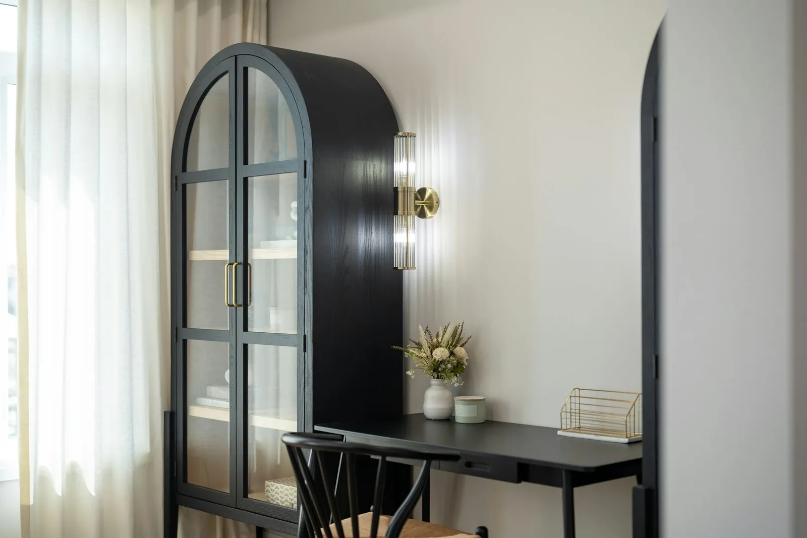 Black arched glass cabinet next to a black desk and chair in a bright home office with a wall sconce