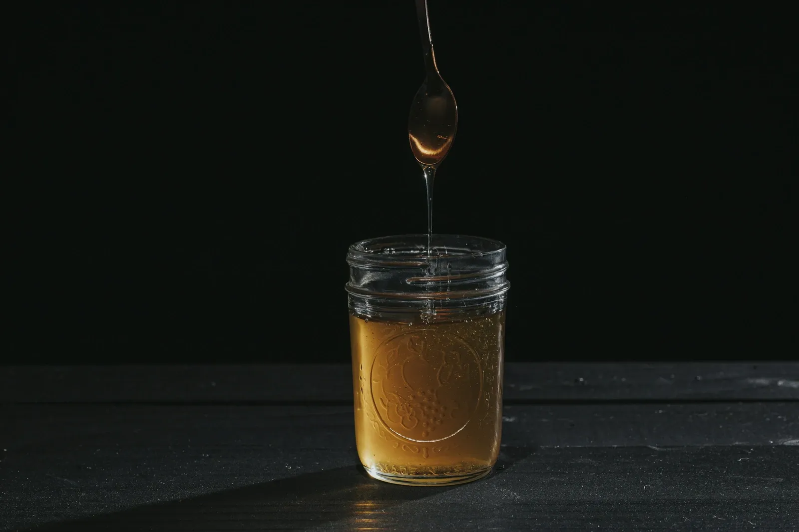 Thick homemade syrup dripping from a spoon into a small glass jar on a dark table
