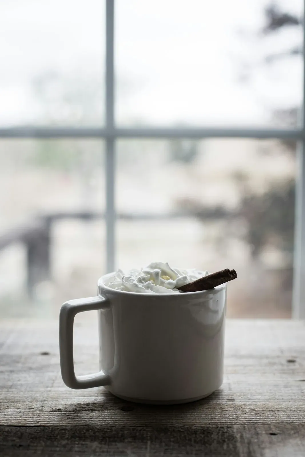 Mug of hot chocolate topped with whipped cream and a cinnamon stick on a windowsill