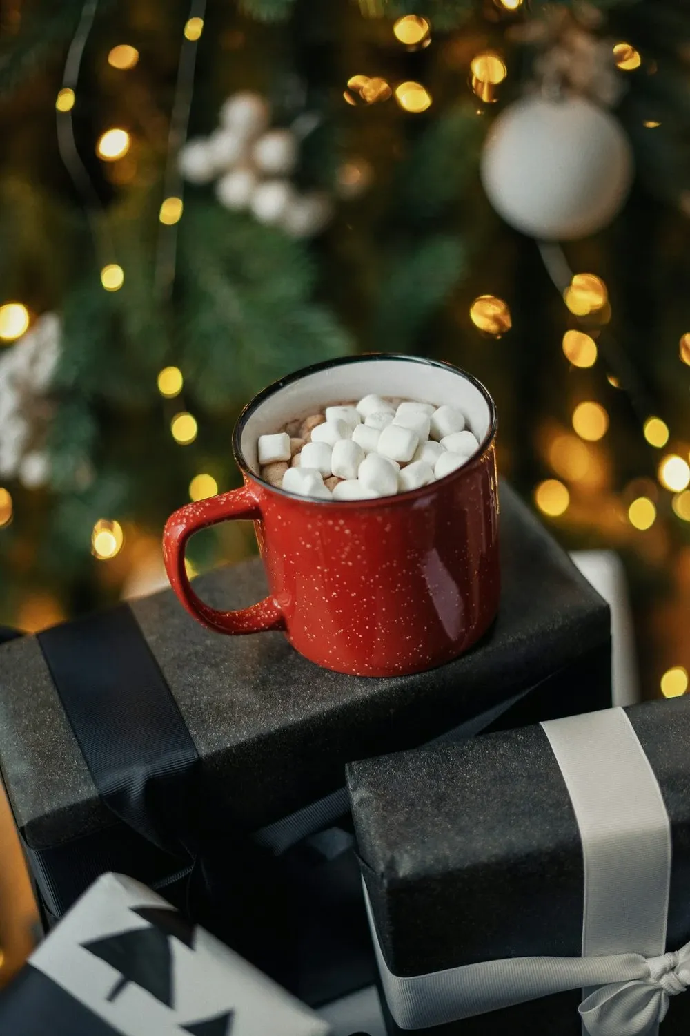Red mug of hot cocoa topped with marshmallows on wrapped gifts in front of Christmas lights