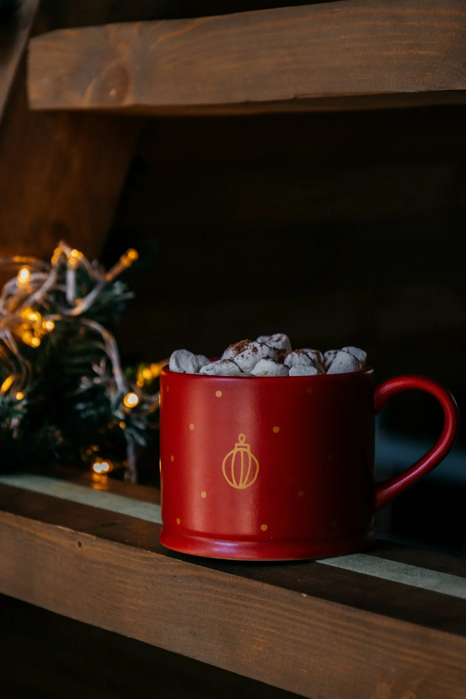 Red mug piled with mini marshmallows on a wooden shelf with warm fairy lights