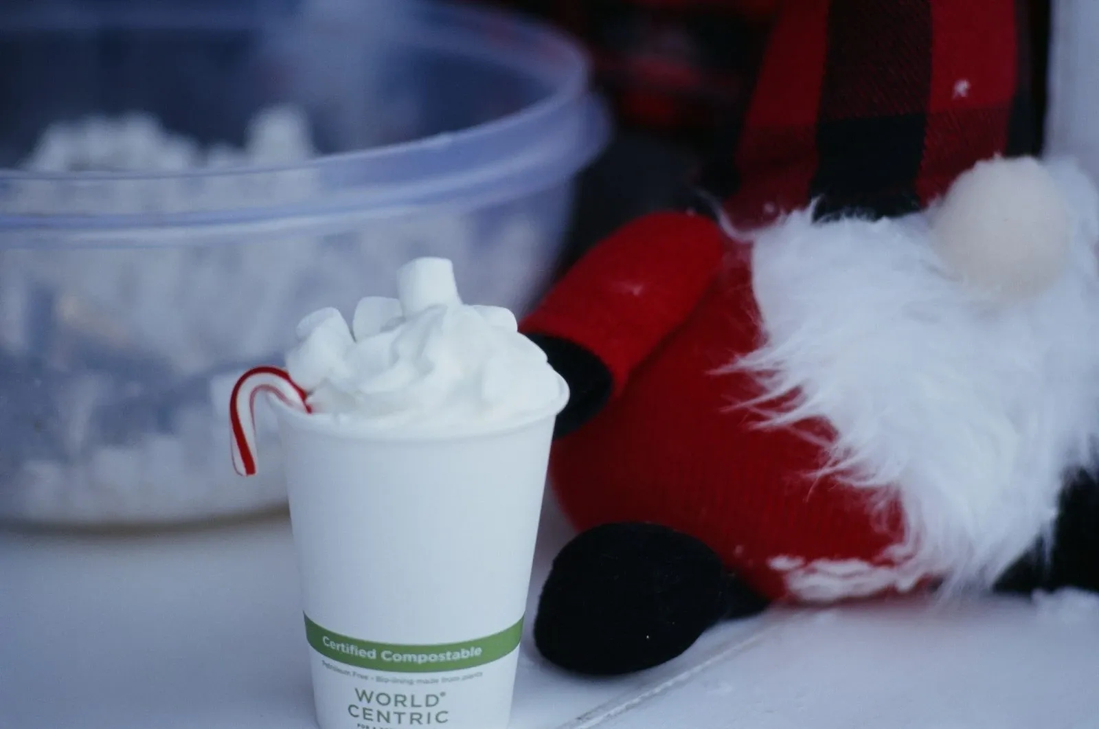 Paper cup of hot cocoa with marshmallows and a candy cane next to a Santa hat and a bowl of marshmallows
