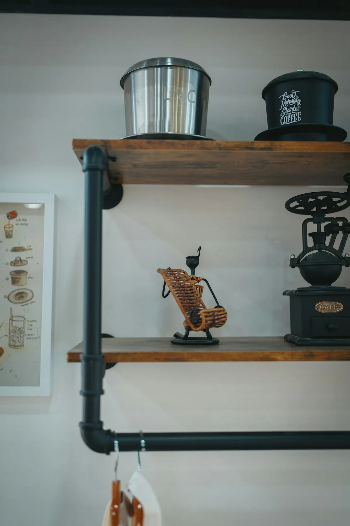 Black pipe and wood wall shelves holding a coffee canister, a hand grinder and mugs hanging from hooks