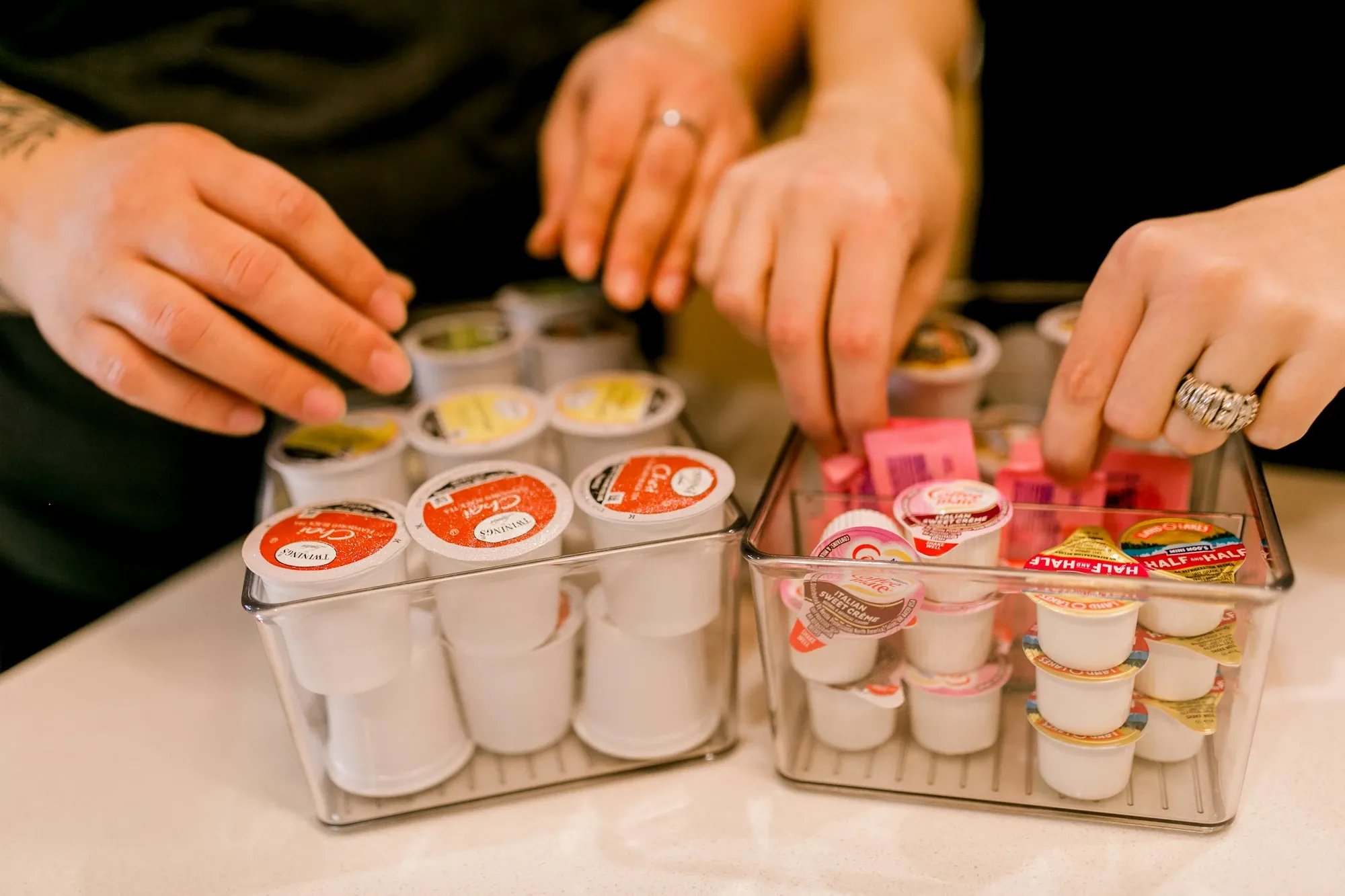 Hands sorting coffee pods and creamer cups into clear acrylic organizers on a white counter