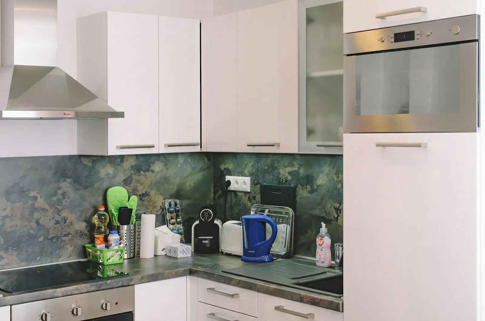 Kitchen corner with a green and gray stone-look backsplash behind a kettle, jars and small appliances under white cabinets