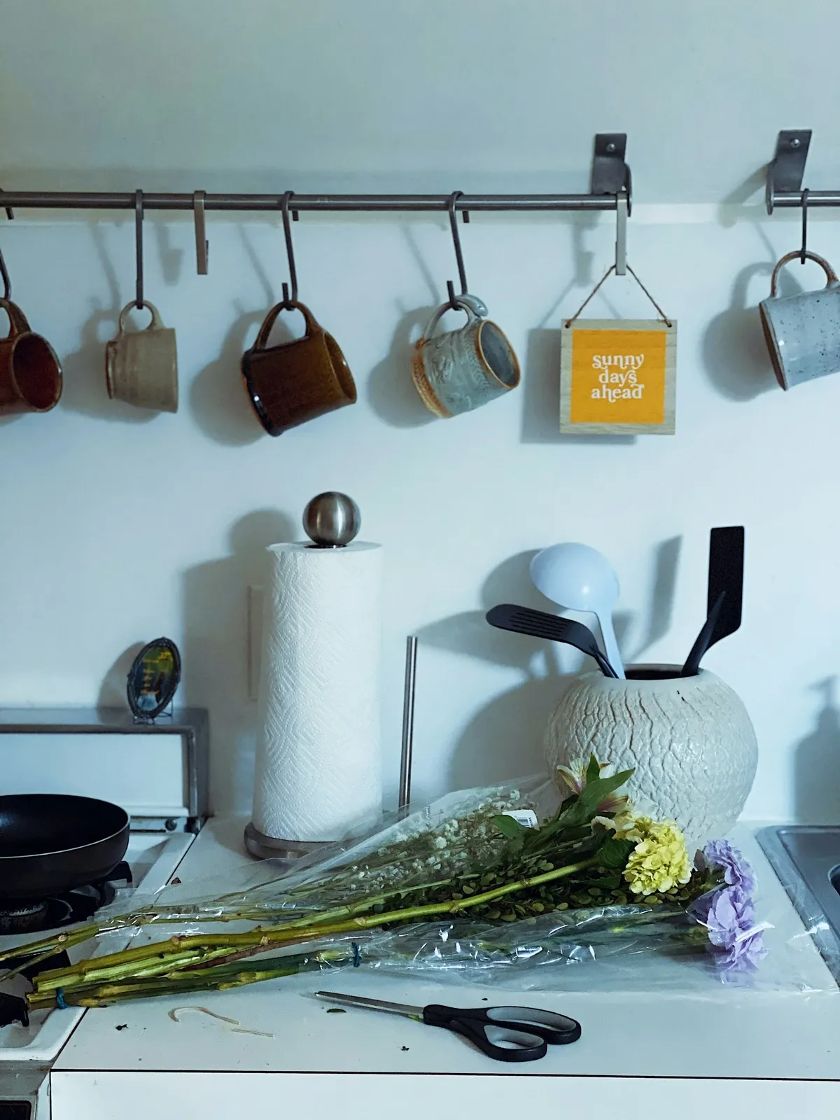 Mugs hanging from S-hooks on a slim steel rail mounted on a white kitchen wall above the counter