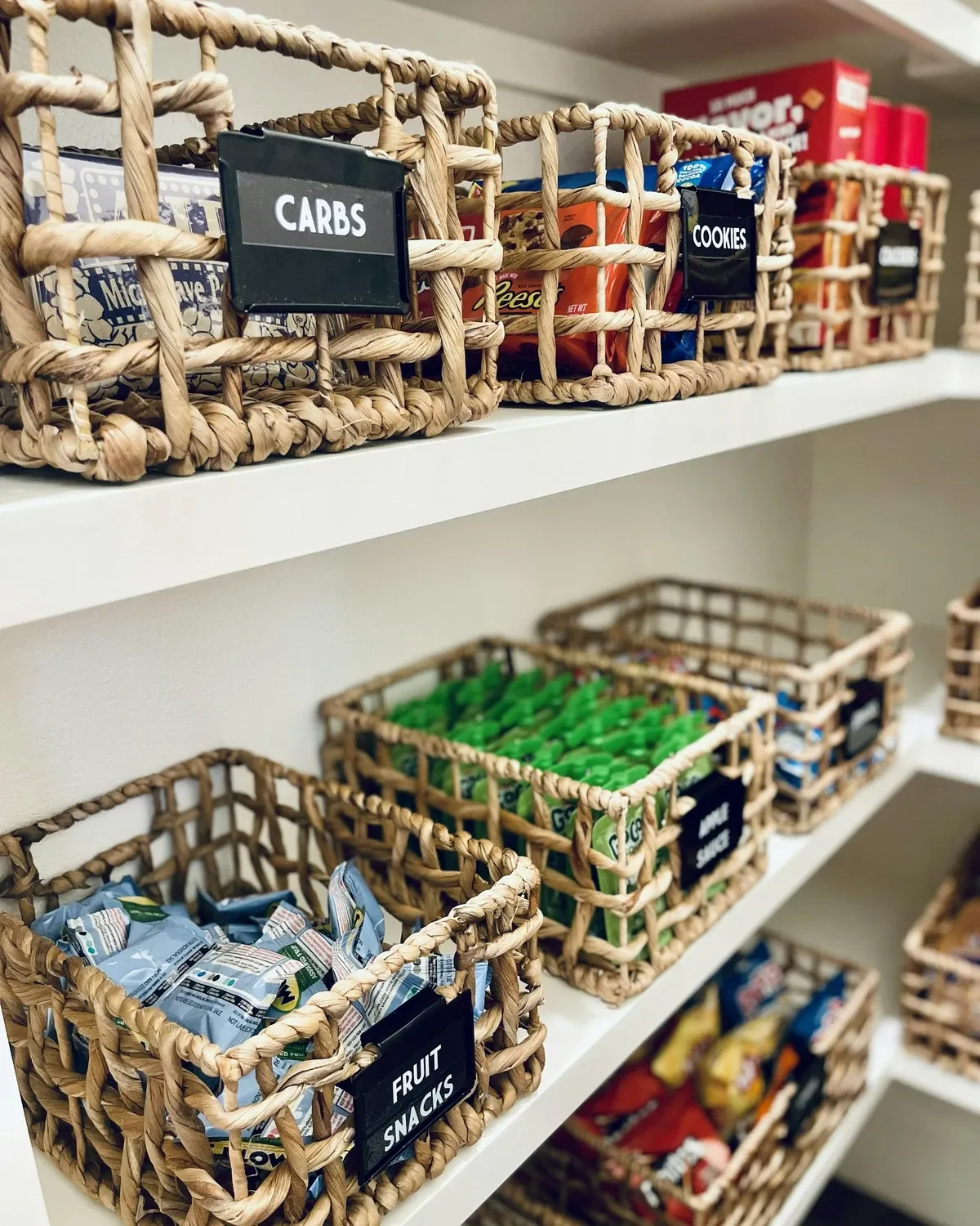 Woven baskets with black labels holding snacks and packets on white pantry shelves