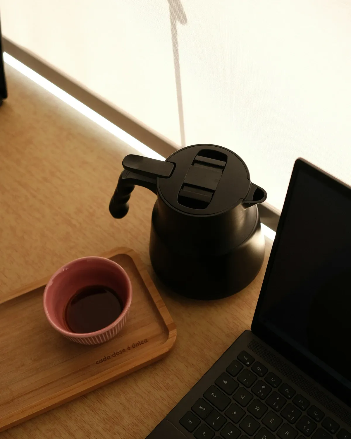 Black electric kettle and a cup of coffee on a wood tray next to an open laptop on a sunny desk