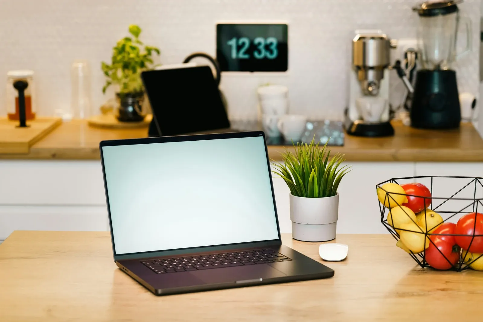 Laptop and a potted plant on a light wood kitchen counter, with an espresso machine and a fruit bowl at the far end