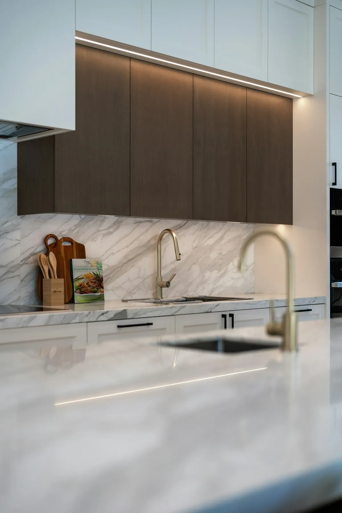 Marble backsplash and counter under dark wood upper cabinets lit from below, with a brass faucet and a small wood tray