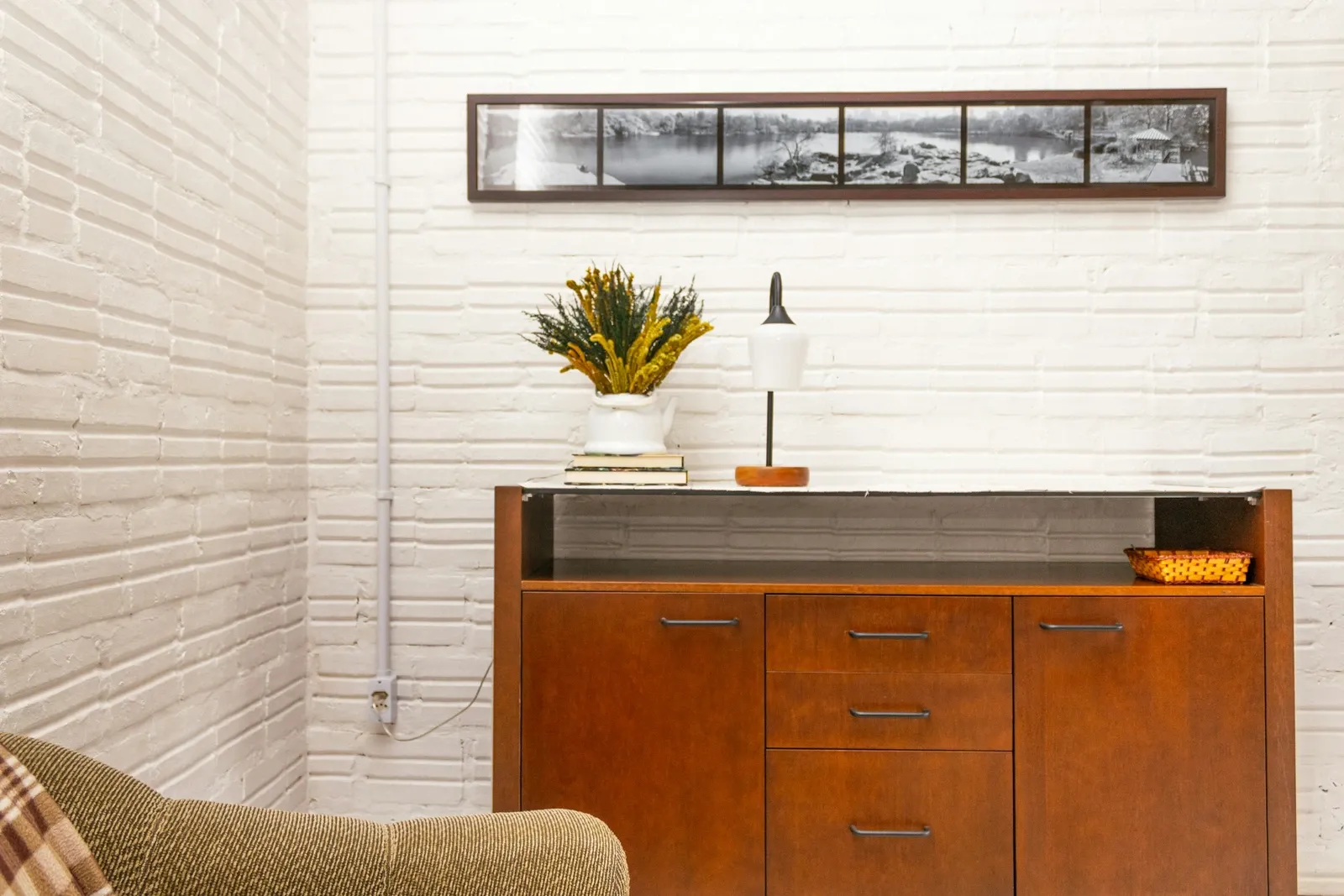 Mid-century wood credenza with sliding doors under a long framed print, with a lamp and a small plant on top