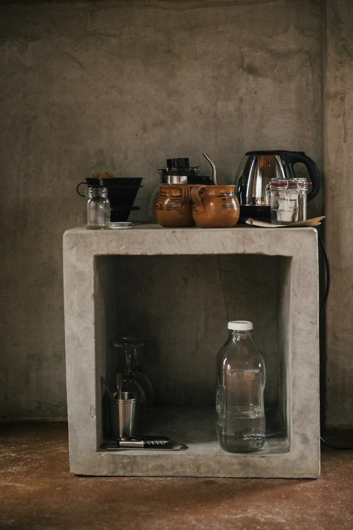 Minimal concrete cube shelf with a coffee pot, a kettle and mugs on top against a gray wall