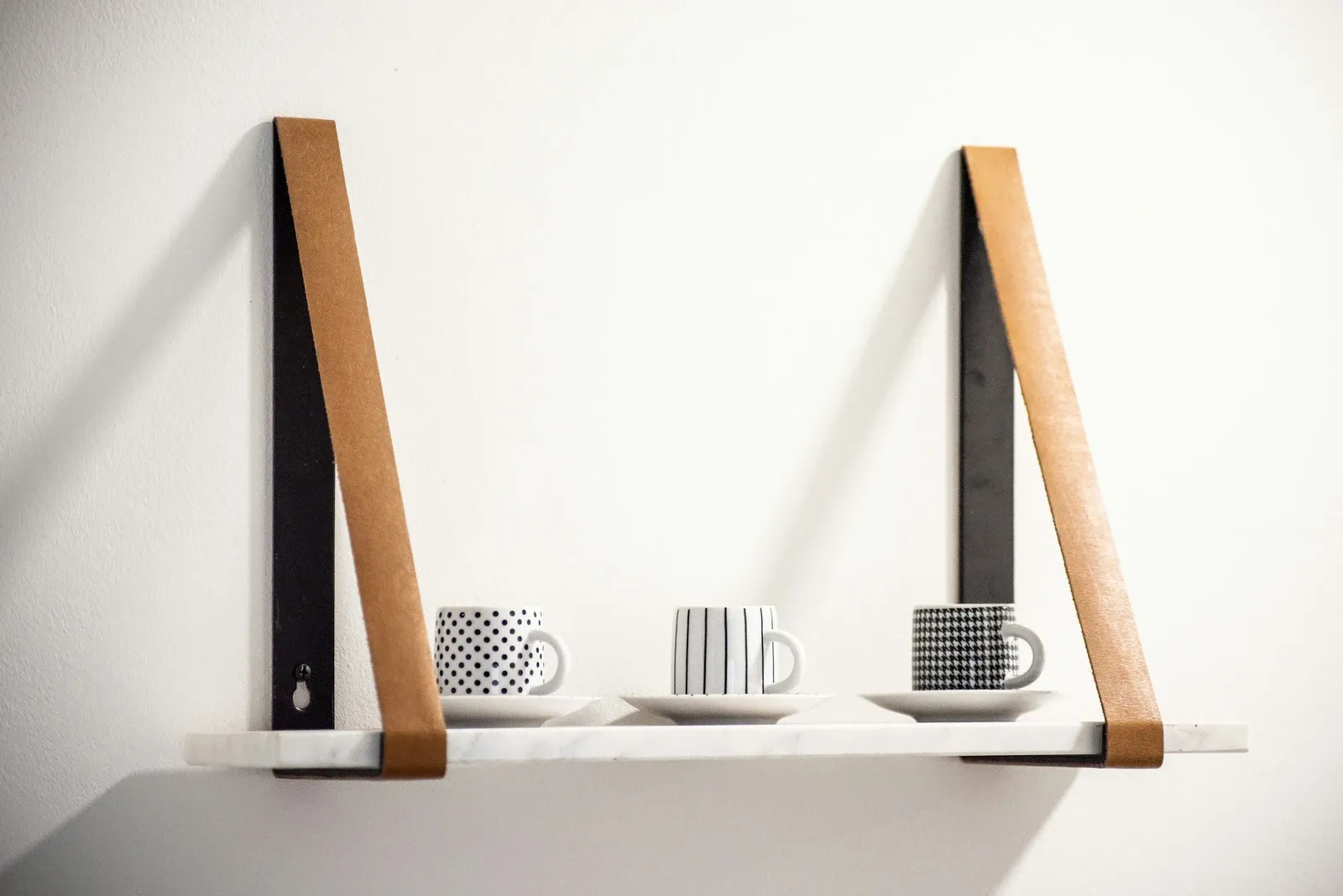 White shelf hung from two brown straps holding three espresso cups on saucers against a white wall