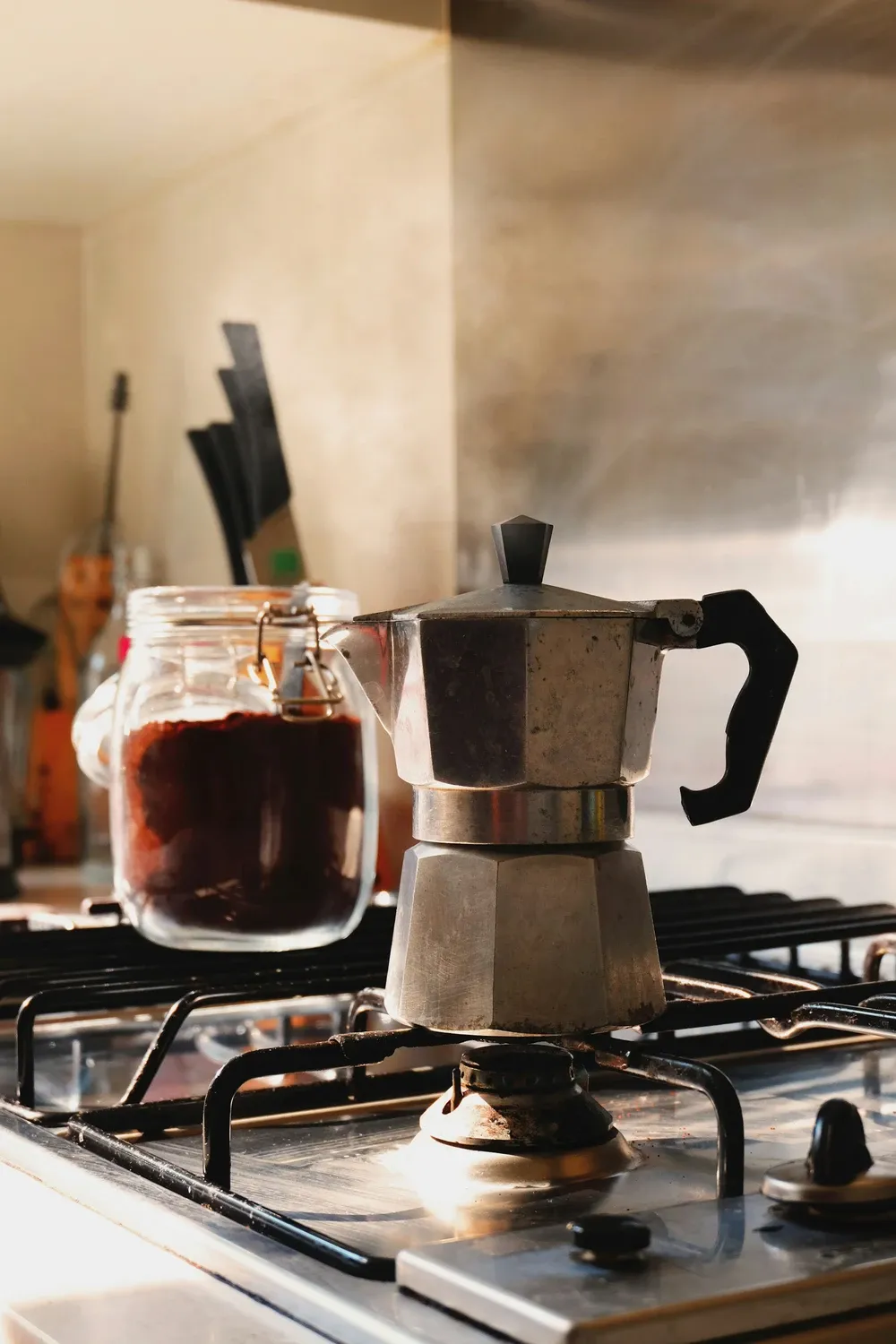Aluminum moka pot on a gas burner beside a glass jar of ground coffee in a home kitchen